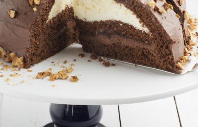 Fotografia em tons de marrom em uma bancada de madeira clara, um suporte de bolo branco com pé preto e uma doce de chocolate com recheio de creme branco e de chocolate.