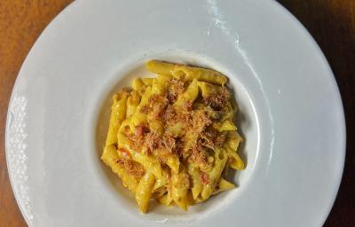 Fotografia vista de cima de um prato de vidro branco com bordas grossas e fundo pequeno sobre uma mesa de madeira. Dentro do prato tem uma receita de penne cremoso com farofa de bacon.
