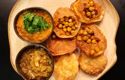 Fotografia vista de cima de seis unidades de pani puri com grão de bico Crocante, e ao lado tem dois recipientes pequenos, um com chutney de cebola e o outro com dahl de lentilha. A receita está em um prato de vidro branco, sobre uma mesa marrom.
