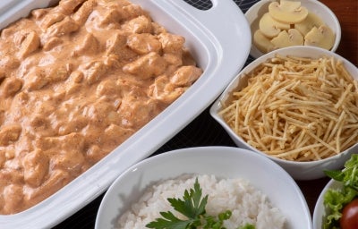 Fotografia de uam travessa branca retangular com o strogonoff de frango, ao redor tem três recipientes. Em cada um tem o champignon fatiado, batata palha e arroz branco. Fotografia de uam travessa branca retangular com o strogonoff de frango, ao redor tem três recipientes. Em cada um tem o champignon fatiado, batata palha e arroz branco.