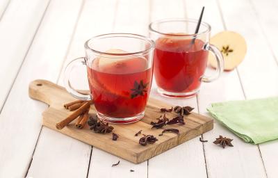 Fotografia em tons de verde em uma bancada de madeira branca, um paninho verde, uma tábua de madeira, duas xícaras de vidro com o chá de hibisco com maçã dentro deles. Ao lado, paus de canela, cravos e anis-estrelado. Fotografia em tons de verde em uma bancada de madeira branca, um paninho verde, uma tábua de madeira, duas xícaras de vidro com o chá de hibisco com maçã dentro deles. Ao lado, paus de canela, cravos e anis-estrelado.