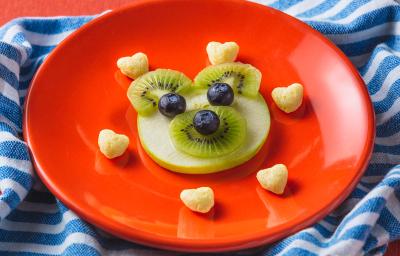 Fotografia em tons de laranja em uma bancada com paninho com listras azuis. Ao centro, um prato laranja redondo com um ursinho feito de maçã, kiwi e mirtilo e acompanhado por biscoitinhos em formato de coração. Ao lado, uma colher laranja e uma embalagem. Fotografia em tons de laranja em uma bancada com paninho com listras azuis. Ao centro, um prato laranja redondo com um ursinho feito de maçã, kiwi e mirtilo e acompanhado por biscoitinhos em formato de coração. Ao lado, uma colher laranja e uma embalagem.