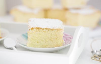 Fotografia em tons claros de um bolo de massa de pão de ló com açúcar de confeiteiro em cima, que está sobre um prato pequeno branco, com desenhos em rosa e azul. Ao fundo, mais pedaços do bolo, e ao lado do prato, colheres de café empilhadas. Fotografia em tons claros de um bolo de massa de pão de ló com açúcar de confeiteiro em cima, que está sobre um prato pequeno branco, com desenhos em rosa e azul. Ao fundo, mais pedaços do bolo, e ao lado do prato, colheres de café empilhadas.