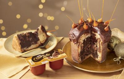 Fotografia em tons de amarelo em uma bancada de madeira clara, um pano amarelo e decoração natalina de fundo. Ao centro, um prato dourado com o panettone de avelã recheado sem um pedaço. Ao lado, um prato com a fatia do panettone.