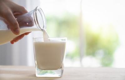 Fotografia de uma mão colocando uma bebida de iogurte com leite e cereal em um copo de vidro, que está sobre uma mesa de madeira em tom claro. Fotografia de uma mão colocando uma bebida de iogurte com leite e cereal em um copo de vidro, que está sobre uma mesa de madeira em tom claro.