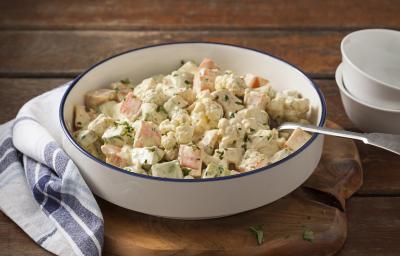 Fotografia em tons de marrom, branco e azul de uma bancada de madeira marrom com uma tábua de madeira, sobre ela um paninho branco xadrez com um recipiente redondo com a salada de legumes e uma colher. Ao lado dois recipientes brancos. Fotografia em tons de marrom, branco e azul de uma bancada de madeira marrom com uma tábua de madeira, sobre ela um paninho branco xadrez com um recipiente redondo com a salada de legumes e uma colher. Ao lado dois recipientes brancos.