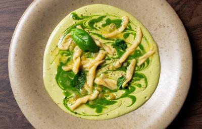 Fotografia em tons de cinza com um prato cinza e fundo ao centro. Dentro do prato existe uma receita de macarrão trofie com molho de abobrinha e ervilhas coberto com pesto de ervas.