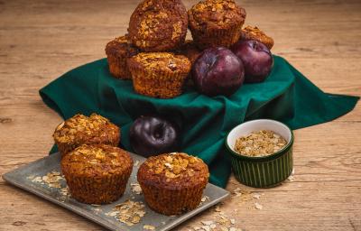 Fotografia em tons de verde em uma bancada de madeira clara, um pano verde, um recipiente retangular com os muffins de ameixa e cereais em cima. Ao lado, um potinho com cereais. Ao fundo, um suporte com o pano verde e mais muffins e ameixas.