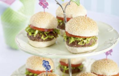 Fotografia em tons de verde em uma mesa branca com um pedestal enfeitado com borboletas de papel de pratos brancos com os hamburguinhos colocados nele. Ao fundo, um copo com guardanapos de papel verde e rosa.