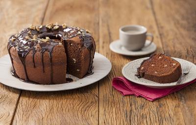 Fotografia em tons de marrom em uma mesa de madeira com um prato branco grande e o bolo de chocolate e nozes e ao lado um prato branco pequeno com uma fatia do bolo. Ao fundo, um pires e uma xícara branca. Fotografia em tons de marrom em uma mesa de madeira com um prato branco grande e o bolo de chocolate e nozes e ao lado um prato branco pequeno com uma fatia do bolo. Ao fundo, um pires e uma xícara branca.