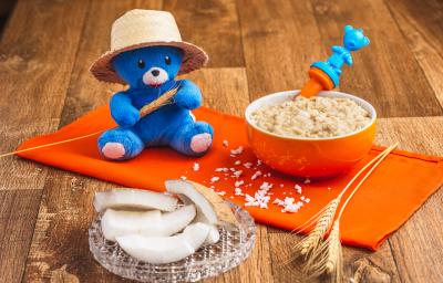 Fotografia em tons de laranja e azul de uma bancada de madeira com um paninho laranja e um pote laranja com a papinha. Na frente, fatias de coco seco decorando e uma pelúcia azul com chapéu de palha.
