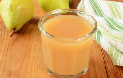 Fotografia em tons de marrom, laranja, branco e verde de uma bancada de madeira marrom vista de cima, contém um copo transparente com suco de pera e maçã. Ao fundo duas peras e ao lado um pano branco e verde