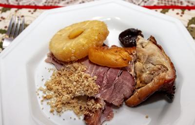 Fotografia vista de cima de um corte de carne, duas fatias de lombo, uma fatia de abacaxi assado e duas frutas assadas em um prato branco. Ao lado do prato, um garfo que está sobre uma toalha de mesa desenhada, na cor branco e verde. Fotografia vista de cima de um corte de carne, duas fatias de lombo, uma fatia de abacaxi assado e duas frutas assadas em um prato branco. Ao lado do prato, um garfo que está sobre uma toalha de mesa desenhada, na cor branco e verde.