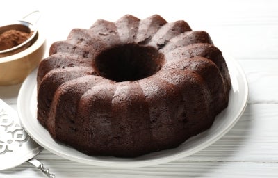 Em cima de uma mesa, bolo de chocolate com aparência escura e densa com úmida e rica em cacau. A textura da superfície é rústica e com coloração marrom intensa. Esse bolo ainda está inteiro, sem cortes ou fatias separadas.