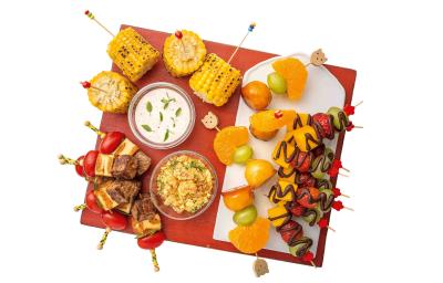 Fotografia em tons de vermelho em uma bancada branca com fundo branco, uma tábua vermelha com vários espetinhos de legumes, carne, frutas com chocolate.