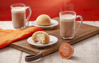 fotografia em tons de laranja e marrom de uma bancada cinza vista de frente, contém uma tábua de madeira com dois pratinhos redondos e brancos com pão de batata com requeijão em cada um, uma xicara transparente com mochaccino canela e um pano laranja.