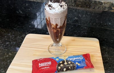 Foto da receita de Milkshake Cookies and Cream, servido em um copo alto decorado com calda de chocolate, com chantilly em cima, sobre uma tábua de madeira com um pacote de Cookies Negresco