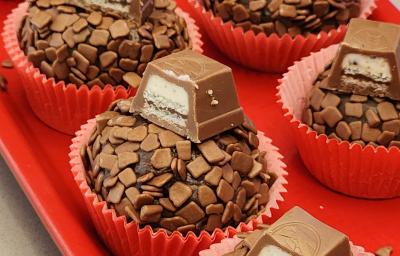 Fotografia de brigadeiros cuidadosamente enrolados em granulado e decorados com pedaços de KitKat, apresentados em forminhas vermelhas sobre um prato da mesma cor.