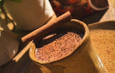 Fotografia em tom claro sob o sol de um prato grande e raso de tom marrom sobre uma mesa de madeira. Apoiado no prato tem um copo de vidro no memso tom com chocolate quente e um pau de cnaela na borda do copo. Fotografia em tom claro sob o sol de um prato grande e raso de tom marrom sobre uma mesa de madeira. Apoiado no prato tem um copo de vidro no memso tom com chocolate quente e um pau de cnaela na borda do copo.