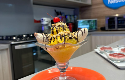 Fotografia de uma taça de vidro com banana split com calda de chocolate, farofa de castanha e sorvete de abacate.