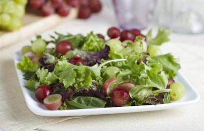 Fotografia de um refratário branco quadrado raso com uma salada de alface, agrião salsão e uva itália com molho de iogurte. O recipiente está apoiado em uma toalha de mesa em tom de bege. Ao fundo tem dois cachos de uva, sendo uvas verdes e uvas roxas.