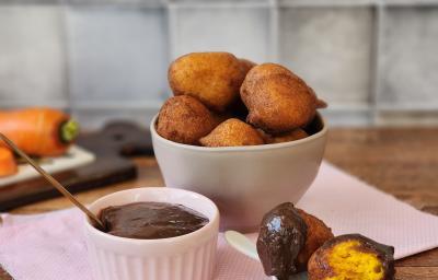 Foto da receita de bolinho de chuva servida em diversas porções em um bowl de cerâmica bege sobre um paninho rosa com um recipiente com a calda a frente. Ao fundo há uma tábua com cenouras em cima de uma mesa de madeira Foto da receita de bolinho de chuva servida em diversas porções em um bowl de cerâmica bege sobre um paninho rosa com um recipiente com a calda a frente. Ao fundo há uma tábua com cenouras em cima de uma mesa de madeira