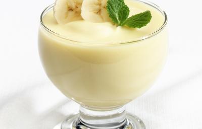 Fotografia de um flan de leite MOÇA e banana dentro de um recipiente de vidro com rodelas de banana e folhas de hortelã. A sobremesa está sobre uma mesa branca.