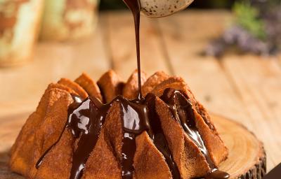 Fotografia em tons de marrom em uma bancada de madeira e ao centro uma tábua de tronco de árvore com o bolo de cenoura em cima e uma mão segurando um potinho despejando a calda de chocolate por cima do bolo. Fotografia em tons de marrom em uma bancada de madeira e ao centro uma tábua de tronco de árvore com o bolo de cenoura em cima e uma mão segurando um potinho despejando a calda de chocolate por cima do bolo.