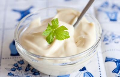 Imagem de um pequeno recipiente redondo de vidro contendo um creme branco. Uma folha de salsinha decora o centro do creme. O Recipiente está disposto sobre uma toalha em tons de branco e azul Imagem de um pequeno recipiente redondo de vidro contendo um creme branco. Uma folha de salsinha decora o centro do creme. O Recipiente está disposto sobre uma toalha em tons de branco e azul