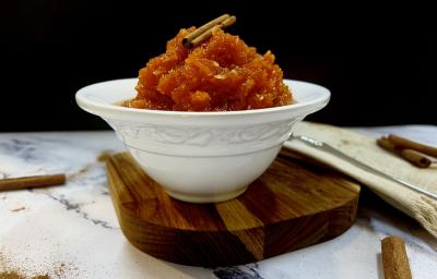 Foto da receita do doce de abóbora com coco servida em um bowl de porcelana branco e com um pau de canela decorativo em cima. O bowl está disposto em cima de uma tábua de madeira, esta em cima de uma mesa de mármore com mais paus de canela decorativos.