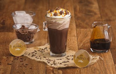 Fotografia em tons de marrom em uma bancada de madeira com um copo de vidro grande e a bebida de chocolate Alpino dentro. Ao fundo, um pote com chantilly, outro com a calda de caramelo e outro com gotas de chocolate e as cápsulas Dolce Gusto Alpino. Fotografia em tons de marrom em uma bancada de madeira com um copo de vidro grande e a bebida de chocolate Alpino dentro. Ao fundo, um pote com chantilly, outro com a calda de caramelo e outro com gotas de chocolate e as cápsulas Dolce Gusto Alpino.