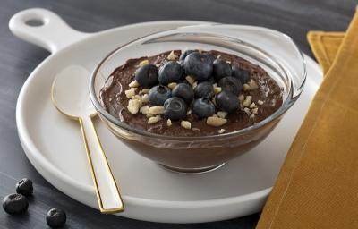 Foto em tons escuros da receita de mousse de chocolate com mirtilo servida em uma taça de vidro sobre uma frigideira de porcelana branca com uma colher branca ao lado