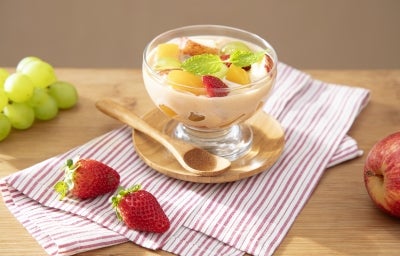 Fotografia em tons claros de um recipiente pequeno decorativo com as frutas e o creme sobre um apoio circular de madeira pequeno ao lado de uma colher pequena de madeira. Fotografia em tons claros de um recipiente pequeno decorativo com as frutas e o creme sobre um apoio circular de madeira pequeno ao lado de uma colher pequena de madeira.
