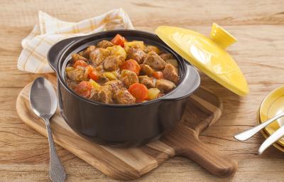 Fotografia em tons de amarelo em uma bancada de madeira clara, um pano amarelo com listras, uma tábua de madeira, uma panela preta de ferro com o picadinho de carne suína com legumes dentro e uma tampa amarela ao lado.