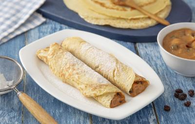 Foto da Receita de Panqueca de Doce de Leite. Observa-se um prato quadrado branco com duas panquecas em cima polvilhadas com açúcar. Do lado esquerdo, tem-se uma peneira com o açúcar e, do direito, as massas de panqueca inteiras. Foto da Receita de Panqueca de Doce de Leite. Observa-se um prato quadrado branco com duas panquecas em cima polvilhadas com açúcar. Do lado esquerdo, tem-se uma peneira com o açúcar e, do direito, as massas de panqueca inteiras.