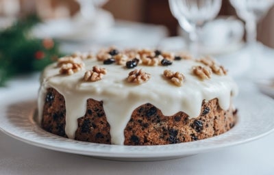 Fotografia de um bolo de frutas decorado com cobertura branca e nozes, apresentado em um prato elegante. O fundo sugere um ambiente festivo e natalino.