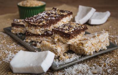 Fotografia em tons de bege em uma bancada de madeira com um jogo americano de palha, um recipiente retangular bege com algumas barrinhas de coco com chocolate em cima dele. Ao lado, pedaços de coco espalhados pela bancada. Fotografia em tons de bege em uma bancada de madeira com um jogo americano de palha, um recipiente retangular bege com algumas barrinhas de coco com chocolate em cima dele. Ao lado, pedaços de coco espalhados pela bancada.