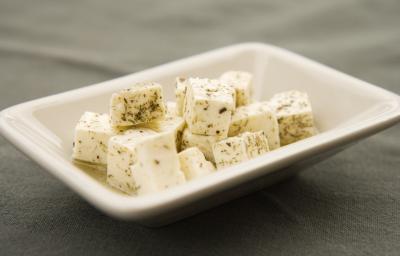 Fotografia de cubos de queijo branco fresco temperados e com azeite dentro de um recipiente pequeno, fundo e quadrado de vidro na cor branco. O aperitivo está sobre uma toalha de mesa preta. Fotografia de cubos de queijo branco fresco temperados e com azeite dentro de um recipiente pequeno, fundo e quadrado de vidro na cor branco. O aperitivo está sobre uma toalha de mesa preta.