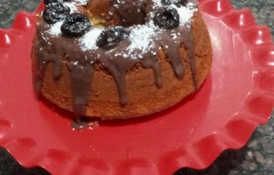 Fotografia de um bolo simples feito na forma de furo central, sobre um apoio grande vermelho decorativo. O bolo está com uma calda de brigadeiro, ameixas e coco ralado por cima. Fotografia de um bolo simples feito na forma de furo central, sobre um apoio grande vermelho decorativo. O bolo está com uma calda de brigadeiro, ameixas e coco ralado por cima.
