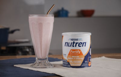 A imagem mostra o frozen de morango servido em um copo de vidro alto estilo de milkshake, com uma lata de nutren fortify ao lado, tudo sobre um pano bege em cima de uma mesa de madeira