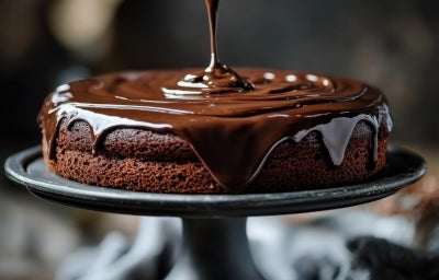 Fotografia de um bolo de chocolate sendo coberto com ganache brilhante e cremoso, posicionado em um suporte escuro. A imagem destaca a textura cremosa do chocolate.
