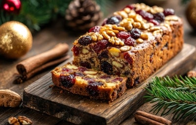 Fotografia de um bolo de frutas festivo, exibido em uma tábua de madeira rústica, repleto de frutas secas e nozes, cercado por elementos natalinos como ramos de pinheiro e canela.