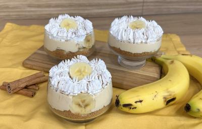 Fotografia em tons de amarelo com um pano amarelo ao centro. Em cima do pano existe uma receita de banoffee feita em pequenos potes de vidro cobertos com chantilly. Ao lado existe duas bananas.