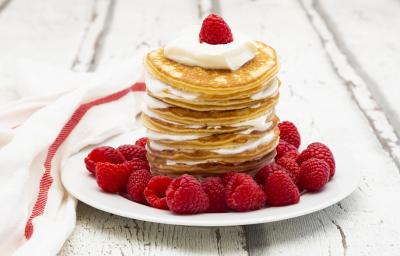Fotografia em tons de branco em uma bancada de madeira de cor branca. Ao centro, um prato branco contendo as pancakes com frutas espalhadas ao redor. Ao lado, um pano branco espalhado. Fotografia em tons de branco em uma bancada de madeira de cor branca. Ao centro, um prato branco contendo as pancakes com frutas espalhadas ao redor. Ao lado, um pano branco espalhado.