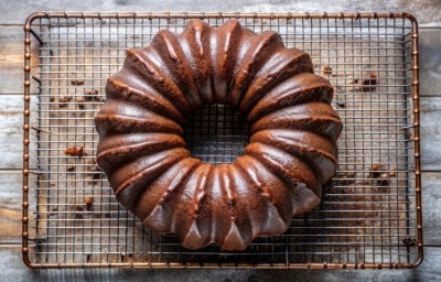 Fotografia de um bolo de chocolate em uma grade de resfriamento, com cor marrom escura e superfície brilhante. O bolo tem um padrão flutuante e está sobre uma superfície de madeira rústica.