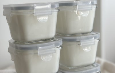 Fotografia em tons de branco com 6 potes ao centro. Dentro de cada pote existe um iogurte natural caseiro.