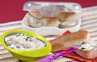 Fotografia em tons de verde, branco e roxo, abaixo potinho com patê cremoso, faca roxa sobre pão aberto ao meio com patê sobre folha de cor salmão, acima recipiente branco com lanches, tudo sobre toalha multicolorida e fundo lilás.