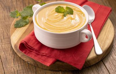 Foto da receita de creme de abóbora com hortelã servida em uma panela de porcelana branca sobre uma tábua de madeira redonda com um pano vermelho entre elas. Para decoração, uma colher prateada ao lado e folhas de hortelã ao fundo. Foto da receita de creme de abóbora com hortelã servida em uma panela de porcelana branca sobre uma tábua de madeira redonda com um pano vermelho entre elas. Para decoração, uma colher prateada ao lado e folhas de hortelã ao fundo.