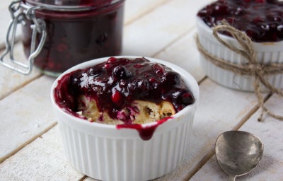Fotografia em tons de roxo em uma bancada de madeira e um pote branco com iogurte e geleia de morango em cima.