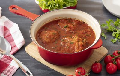 Foto em tons de vermelho da receita de frango com molho de tomate caseiro servida em uma panela vermelha sobre uma tábua de madeira retangular. Ao lado, há tomates e alfaces de decoração, além de um paninho quadriculado branco e vermelho